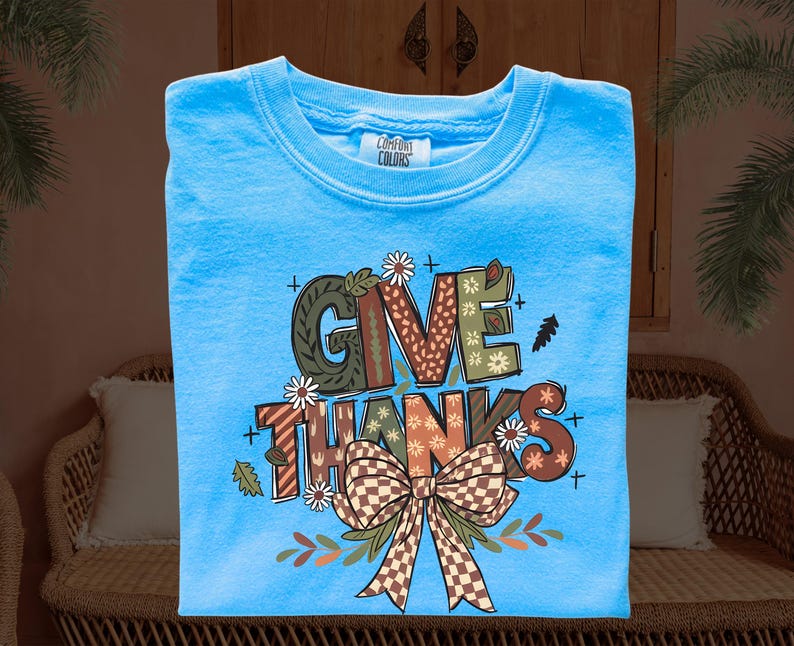 May include: A light blue Comfort Colors t-shirt with the words "GIVE THANKS" in a fall-themed design. The design includes a brown and green plaid bow, leaves, and floral accents. The shirt is a crew neck.