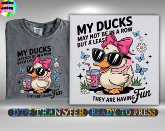 May include: A gray t-shirt and a white canvas print featuring a cartoon duck wearing sunglasses and a pink bow. The text reads "MY DUCKS MAY NOT BE IN A ROW BUT A LEAST THEY ARE HAVING Fun". Butterflies and flowers surround the duck.
