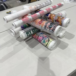 May include: A collection of rolled-up posters, each featuring colorful designs and text. The posters are stacked on a white table, with some displaying sports-related graphics and team names. The overall theme suggests promotional materials or artwork.