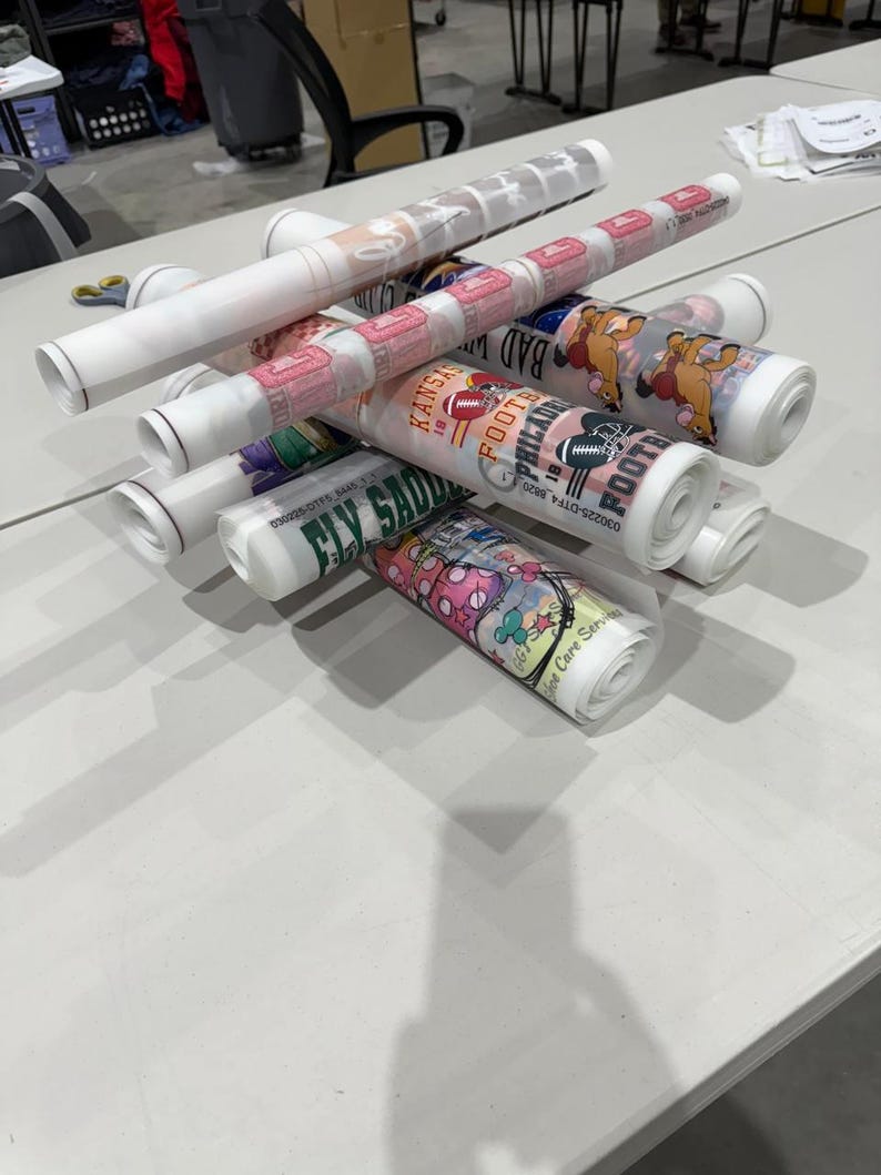 May include: A collection of rolled-up posters, each featuring colorful designs and text. The posters are stacked on a white table, with some displaying sports-related graphics and team names. The overall theme suggests promotional materials or artwork.