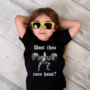 May include: A black t-shirt featuring the phrase "Dost thou even hoist?" above a graphic of a knight lifting weights. The person in the image is wearing yellow sunglasses.