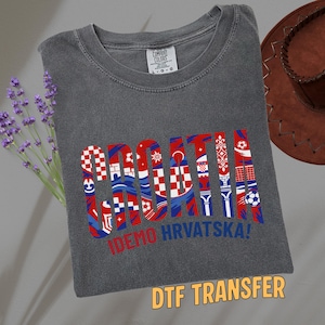 May include: Dark gray t-shirt with the word "CROATIA" in red, white, and blue, with various Croatian flag-inspired designs within the letters. Below the word is the text "IDEMO HRVATSKA!". The shirt also has a "DTF TRANSFER" label.