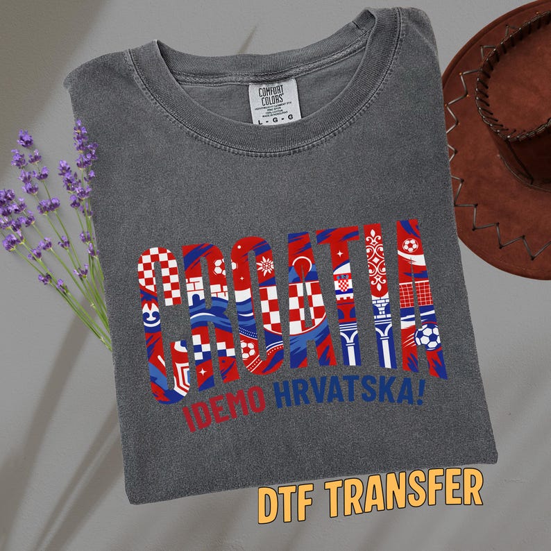 May include: Dark gray t-shirt with the word "CROATIA" in red, white, and blue, with various Croatian flag-inspired designs within the letters. Below the word is the text "IDEMO HRVATSKA!". The shirt also has a "DTF TRANSFER" label.