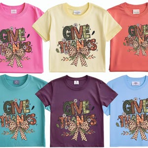 May include: Six short-sleeved t-shirts in various colors: pink, yellow, coral, teal, purple, and light blue. Each shirt features the words "GIVE THANKS" in a decorative font, with a large bow and floral accents. A festive design for the Thanksgiving holiday.
