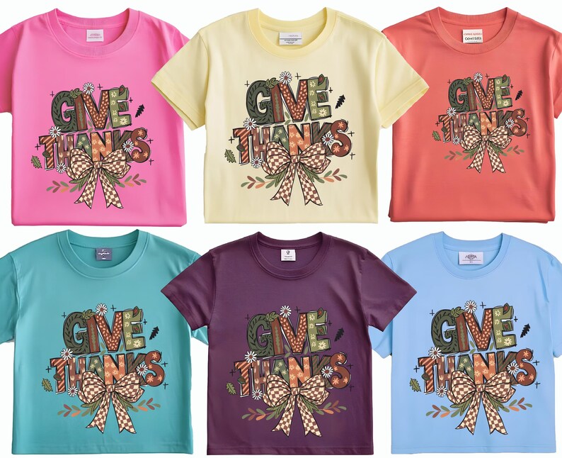 May include: Six short-sleeved t-shirts in various colors: pink, yellow, coral, teal, purple, and light blue. Each shirt features the words "GIVE THANKS" in a decorative font, with a large bow and floral accents. A festive design for the Thanksgiving holiday.