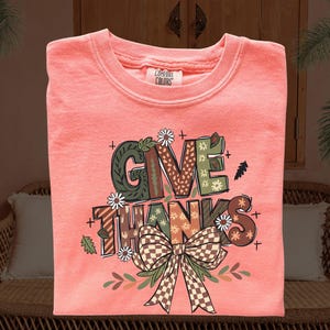 May include: Coral-colored sweatshirt with the words "GIVE THANKS" in a decorative font. The design includes a bow, leaves, and floral accents. The sweatshirt is made by Comfort Colors.