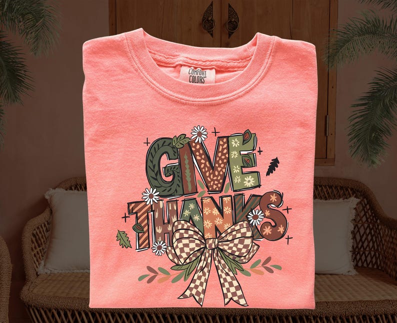 May include: Coral-colored sweatshirt with the words "GIVE THANKS" in a decorative font. The design includes a bow, leaves, and floral accents. The sweatshirt is made by Comfort Colors.