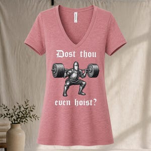 May include: A dusty rose V-neck t-shirt featuring a graphic of a knight lifting a barbell. The text "Dost thou even hoist?" is above and below the knight illustration. The shirt is made of a soft material.