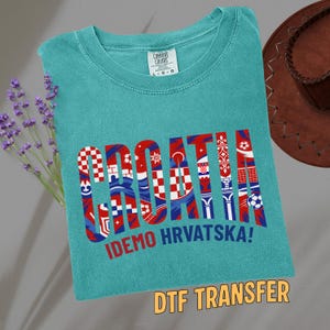 May include: Teal t-shirt with the word "CROATIA" in a red, white, and blue design. The text "IDEMO HRVATSKA!" is below the word. A brown cowboy hat and lavender sprigs are in the background.