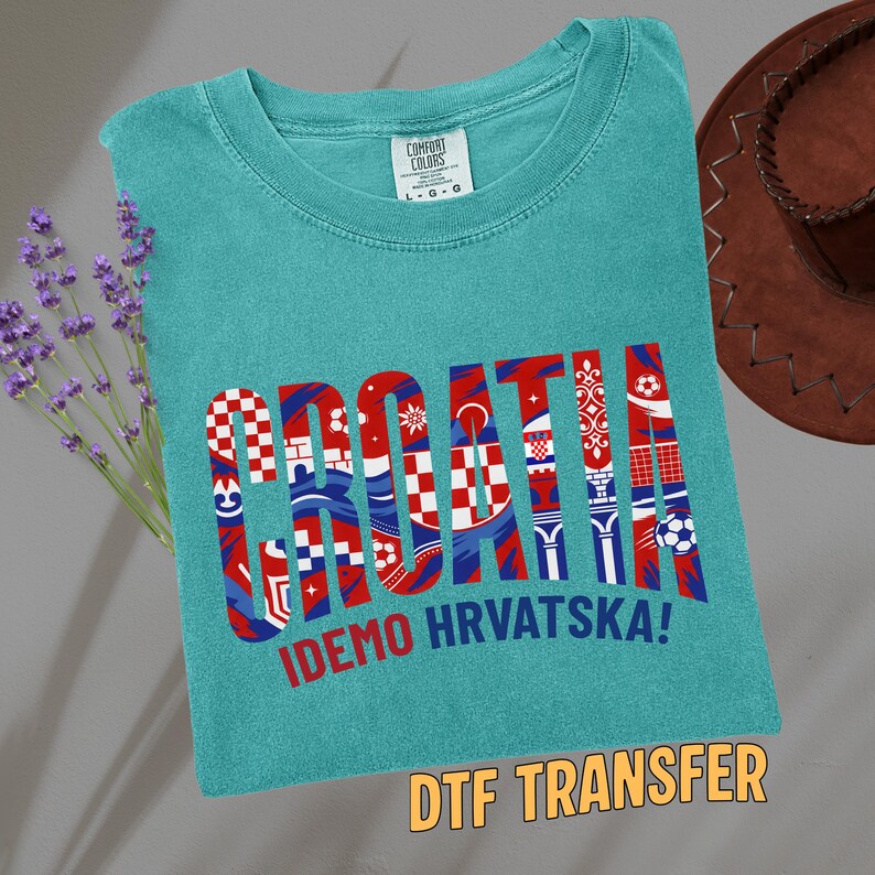 May include: Teal t-shirt with the word "CROATIA" in a red, white, and blue design. The text "IDEMO HRVATSKA!" is below the word. A brown cowboy hat and lavender sprigs are in the background.