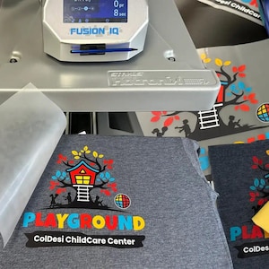 May include: A heat press machine is used to apply a colorful graphic design to a gray t-shirt. The design includes a treehouse, the word "PLAYGROUND", and the text "ColDesi ChildCare Center". A yellow t-shirt is also visible.