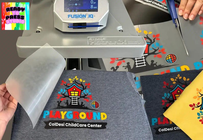 May include: A heat press machine is used to apply a colorful graphic design to a gray t-shirt. The design includes a treehouse, the word "PLAYGROUND", and the text "ColDesi ChildCare Center". A yellow t-shirt is also visible.