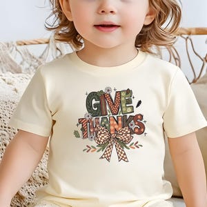 May include: A cream-colored t-shirt featuring the words "GIVE THANKS" in a colorful, decorative font. The design includes a bow and floral accents, perfect for Thanksgiving. The shirt is worn by a young child.