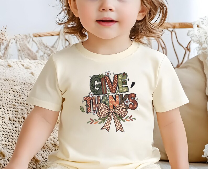 May include: A cream-colored t-shirt featuring the words "GIVE THANKS" in a colorful, decorative font. The design includes a bow and floral accents, perfect for Thanksgiving. The shirt is worn by a young child.