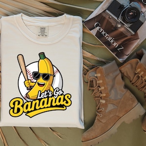 May include: Cream-colored t-shirt featuring a cartoon banana wearing sunglasses and holding a baseball bat, set against a baseball. The text "Let's Go Bananas" is in yellow. The shirt is folded.