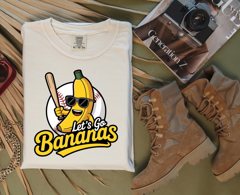 May include: Cream-colored t-shirt featuring a cartoon banana wearing sunglasses and holding a baseball bat, set against a baseball. The text "Let's Go Bananas" is in yellow. The shirt is folded.