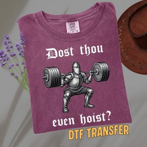 May include: A maroon t-shirt with the text "Dost thou even hoist?" and a graphic of a knight lifting weights. The shirt also has the text "DTF TRANSFER". A cowboy hat and lavender sprigs are in the background.