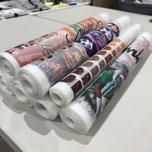 May include: Several rolls of printed fabric are stacked on a white table. The rolls display various colorful designs, including cartoon characters, abstract patterns, and a sports-themed image. The fabric is rolled tightly, showing the printed designs on the outer layer.