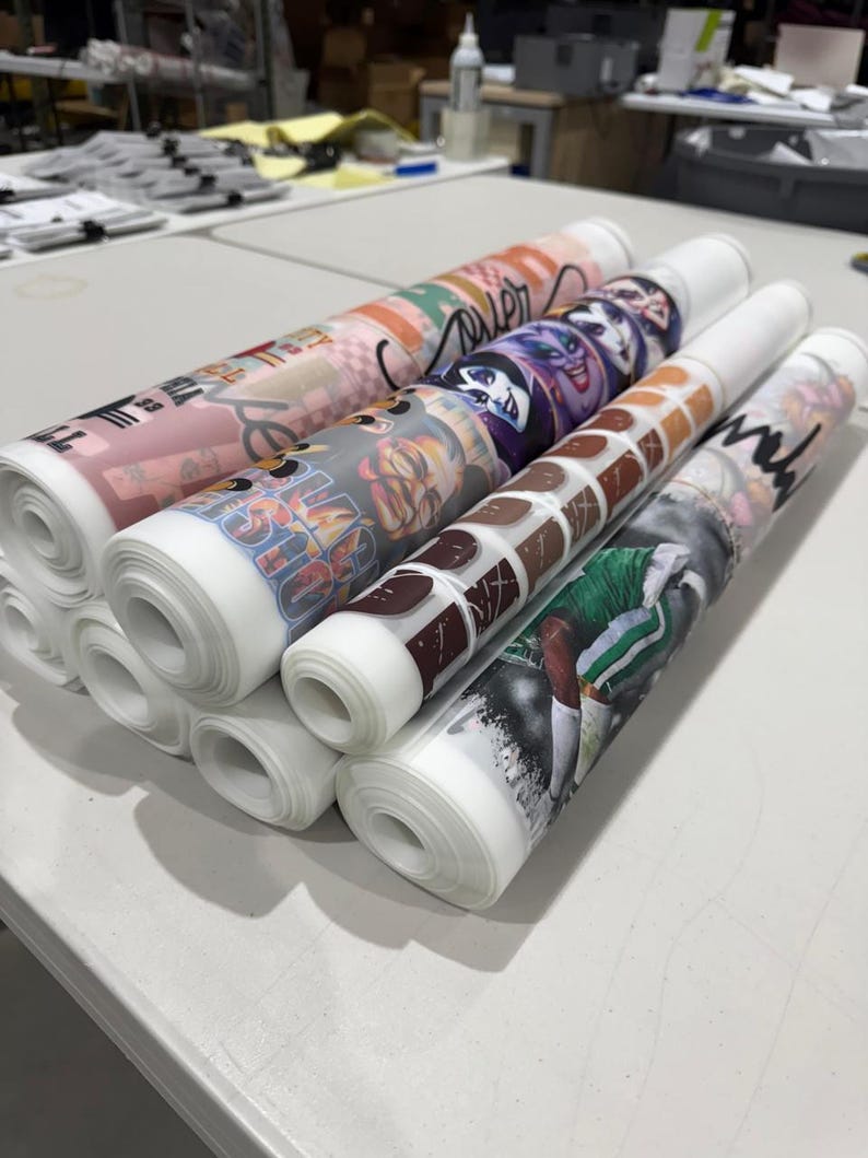 May include: Several rolls of printed fabric are stacked on a white table. The rolls display various colorful designs, including cartoon characters, abstract patterns, and a sports-themed image. The fabric is rolled tightly, showing the printed designs on the outer layer.