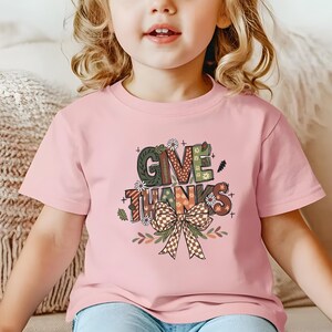 May include: A light pink t-shirt featuring the words "GIVE THANKS" in a colorful, decorative font. The design includes a bow and floral accents, perfect for Thanksgiving. The shirt is worn by a young child.