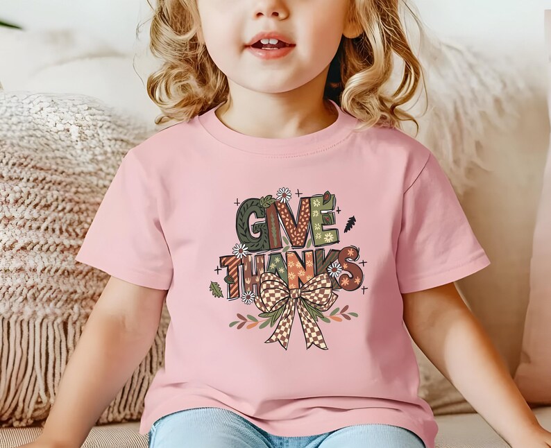 May include: A light pink t-shirt featuring the words "GIVE THANKS" in a colorful, decorative font. The design includes a bow and floral accents, perfect for Thanksgiving. The shirt is worn by a young child.