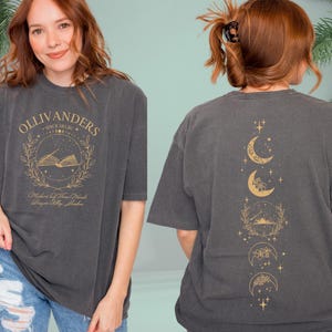 May include: Dark grey t-shirt with gold text and graphics. The front displays "OLLIVANDERS" above an open book in a wreath. The back shows a vertical design of moon phases. The shirt is worn by a person.