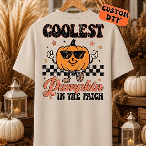 May include: A beige short-sleeve t-shirt featuring a graphic of an orange pumpkin wearing black sunglasses and making peace signs. The text reads "COOLEST" above and "Pumpkin IN THE PATCH" below, with a black and white checkered pattern.