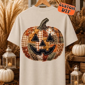 May include: A beige short-sleeve t-shirt featuring a large graphic of a pumpkin made from various fabric patches in orange, brown, and cream tones. The pumpkin has a black stitched face and the text CUSTOM DTF in an orange bubble.