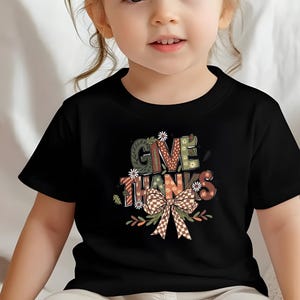 May include: Black t-shirt featuring the words "GIVE THANKS" in a colorful, decorative font. The design includes a bow and floral accents. The shirt is worn by a young child.