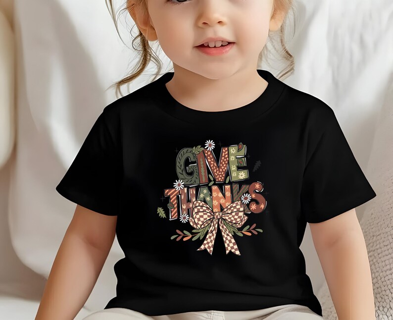 May include: Black t-shirt featuring the words "GIVE THANKS" in a colorful, decorative font. The design includes a bow and floral accents. The shirt is worn by a young child.