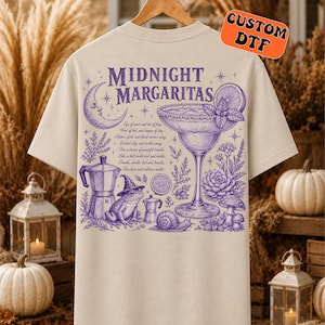 May include: A beige t-shirt featuring a purple graphic on the back with the text "MIDNIGHT MARGARITAS". The design includes a crescent moon, a margarita glass, a frog wearing a witch hat, a snail, mushrooms, and a coffee maker, surrounded by botanical elements and written incantations.