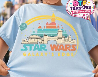 May include: Pale blue t-shirt with a colourful graphic depicting a Star Wars-themed landscape and the text "STAR WARS GALAXY'S EDGE". The design includes a spaceship, a castle, and a rainbow. A "DTF TRANSFER READY TO PRESS" sticker is on the shirt.
