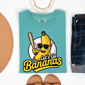 May include: Teal t-shirt with a cartoon banana wearing sunglasses and holding a baseball bat, in front of a baseball. The text "Let's Go Bananas" is below the graphic. The shirt is paired with denim shorts and a brown hat.
