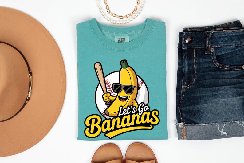 May include: Teal t-shirt with a cartoon banana wearing sunglasses and holding a baseball bat, in front of a baseball. The text "Let's Go Bananas" is below the graphic. The shirt is paired with denim shorts and a brown hat.
