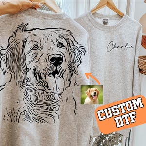 May include: Two light gray sweatshirts. One features a large black line drawing of a dog's face on the back. The other has the name "Charlie" in cursive. A small photo of a golden retriever is included. The orange sign says "CUSTOM DTF".