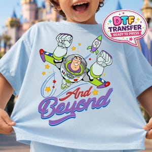 May include: Light blue t-shirt featuring a colorful graphic of Buzz Lightyear in flight, with a rocket and stars. The phrase "And Beyond" is written in a stylized font. A speech bubble with the text "DTF TRANSFER READY TO PRESS" is also visible.