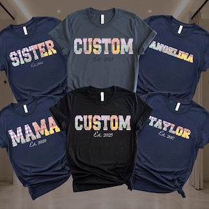 May include: Five short-sleeve t-shirts in navy, charcoal, and black. The shirts feature custom text in a colorful plaid pattern, including the words SISTER, CUSTOM, ANGELINA, MAMA, and TAYLOR, along with established years like Est. 2002, 2025, and 2017.