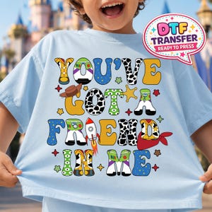 May include: Light blue t-shirt with the phrase "YOU'VE GOT A FRIEND IN ME" in colorful, cartoon-style letters. The design includes a cowboy hat, star, rocket, and other playful elements. A DTF transfer label is in the upper right corner.