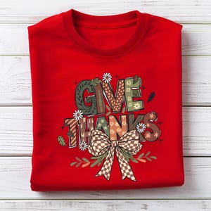 May include: A folded red sweatshirt with the words "GIVE THANKS" in a decorative font. The letters are filled with various patterns, including floral, plaid, and geometric designs. A large bow and floral accents are below the text.
