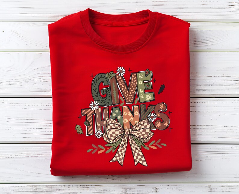 May include: A folded red sweatshirt with the words "GIVE THANKS" in a decorative font. The letters are filled with various patterns, including floral, plaid, and geometric designs. A large bow and floral accents are below the text.