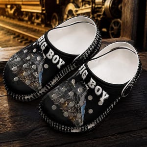 May include: A pair of black clogs featuring a graphic print of a steam locomotive and the text BIG BOY in white lettering. The shoes have a white interior, a rounded toe, and a series of circular ventilation holes across the top surface.