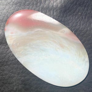 Puede incluir: Una piedra preciosa ovalada y pulida. La sección superior es de un rojo suave, que se mezcla con blanco y azul claro. La piedra tiene una superficie lisa y brillante. Los colores crean un efecto veteado.