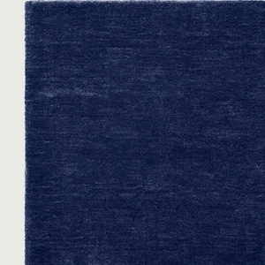 May include: A close-up of a section of a navy blue rug. The rug has a soft, textured surface and a solid color. The edges of the rug are visible, with a white background on the left side.