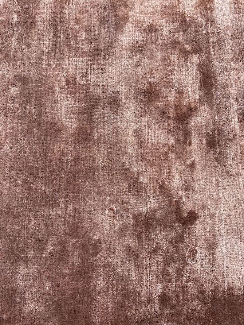 May include: A close-up shot of a dusty rose-colored rug with a textured, distressed appearance. The rug has a soft, velvety feel with variations in color, creating a vintage aesthetic. The rug's surface shows subtle patterns and a worn look.
