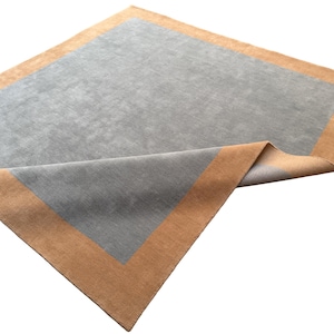 May include: A rectangular area rug featuring a solid light gray center panel framed by a wide, solid tan border. The rug is shown with one corner folded over, revealing the gray underside and the consistent texture of the fabric surface.