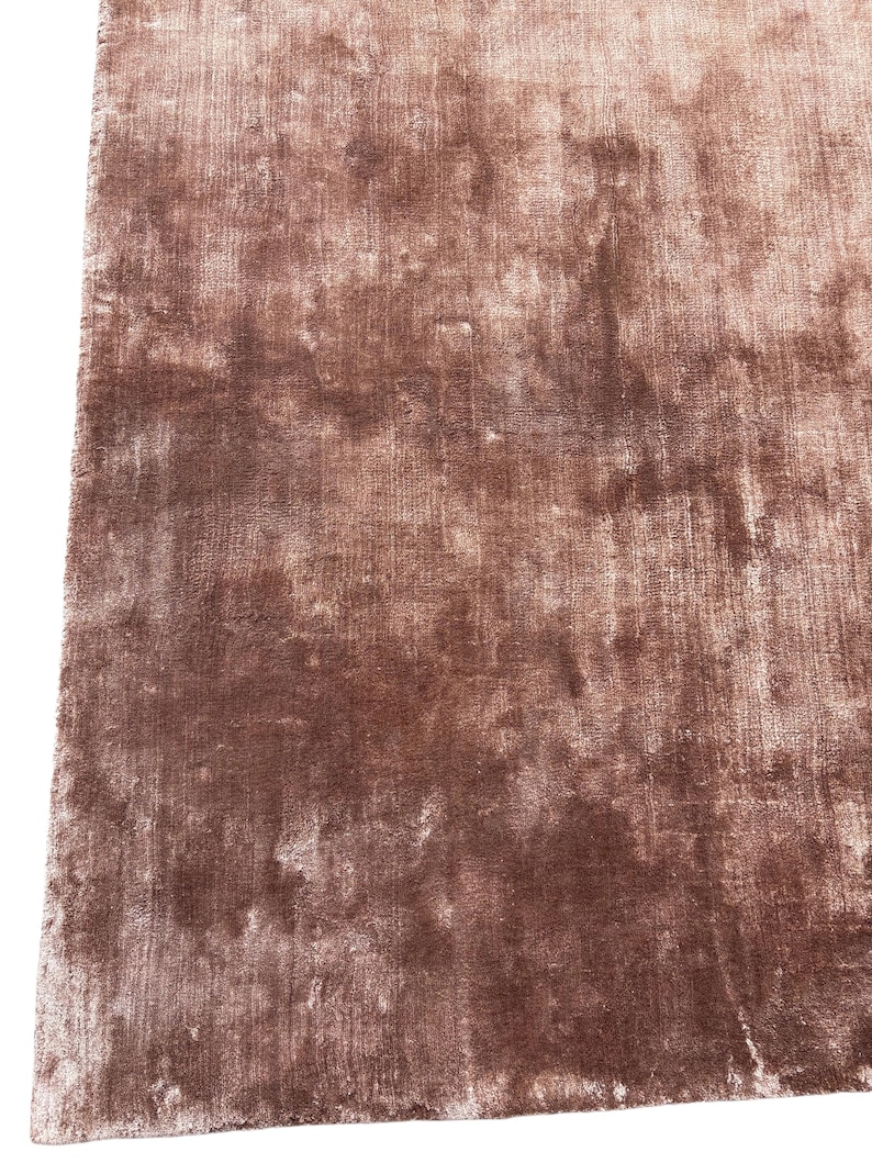 May include: A close-up shot of a rectangular, plush rug in a muted, dusty rose color. The rug has a soft, textured appearance with subtle variations in tone, creating a luxurious feel. The fibers appear dense and slightly reflective, suggesting a high-quality material.