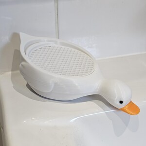 May include: A white, duck-shaped soap dish with an orange beak and black eyes. The dish has a textured, perforated surface and is designed to hold soap. The duck is resting on a white surface.