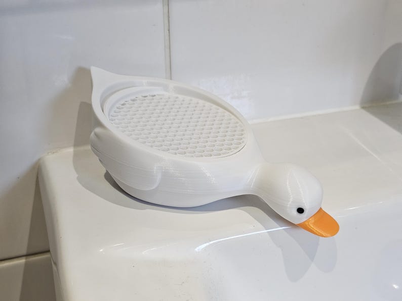 May include: A white, duck-shaped soap dish with an orange beak and black eyes. The dish has a textured, perforated surface and is designed to hold soap. The duck is resting on a white surface.