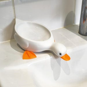 May include: A white duck-shaped soap dish with orange feet and beak. The dish is on a white sink, next to a chrome faucet. The duck's head is angled downward, as if looking at the sink.