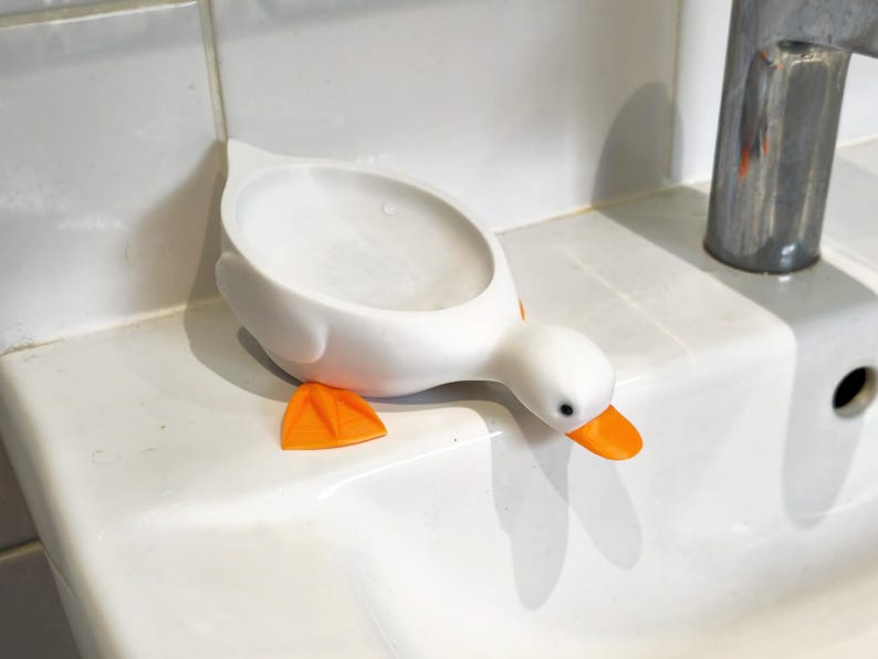 May include: A white duck-shaped soap dish with orange feet and beak. The dish is on a white sink, next to a chrome faucet. The duck's head is angled downward, as if looking at the sink.